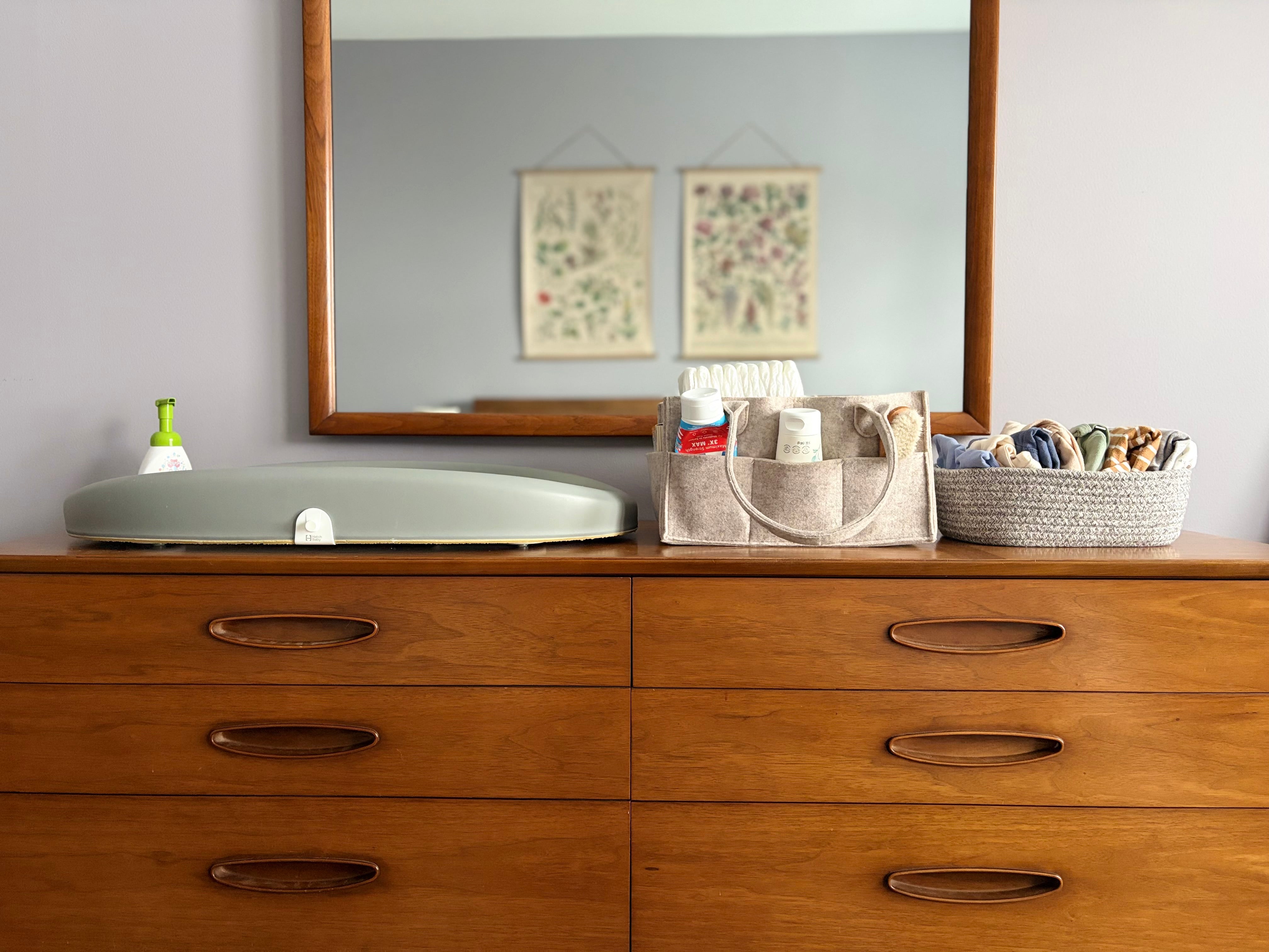 Wooden dresser with mirror, diaper caddy, and a basket of baby clothes in a bedroom.